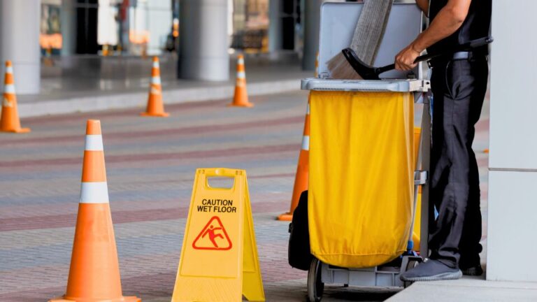 A janitor stands with a cleaning trolley near the entrance of a modern office building, ready to start his cleaning duties.