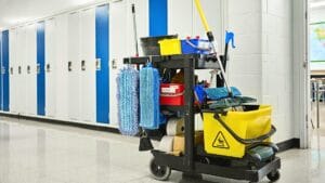 Janitorial cart with cleaning supplies positioned in a school hallway with lockers and bright lighting, ready for custodial use.